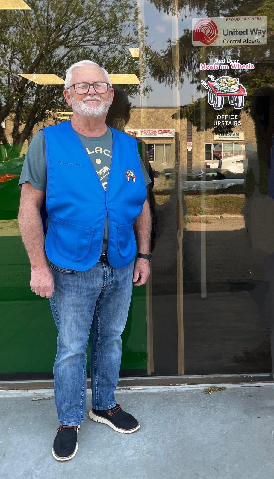 A smiling volunteer in a blue vest outside the Red Deer Meals on Wheels office